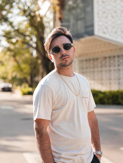 EVIE EYEWEAR MODELO IBIZA oval sunglasses with gold frames and dark lenses, worn by a man in a cream t-shirt.