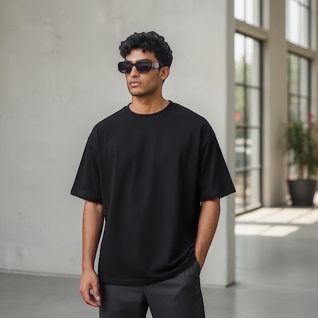 Hombre con gafas de sol EVIE EYEWEAR ORÉA negras, camiseta negra holgada y pantalones grises.